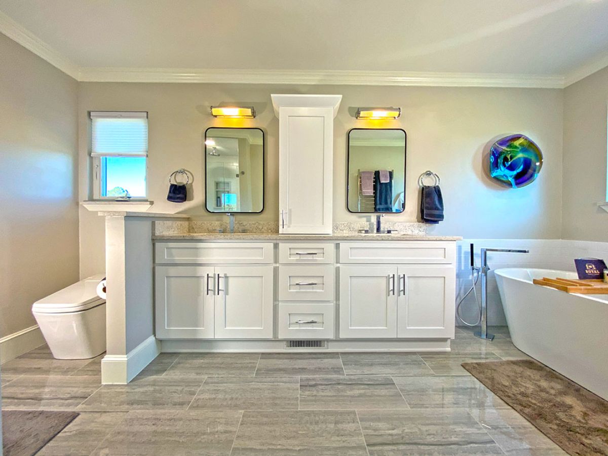 A remodeled bathroom with white two sink cabinets, two mirrors, bidet, and stand-alone tub.